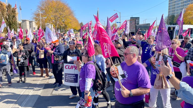 Image of a crowd of people in front of the Toronto Congress Centre. Many wear purple shirts holding red flags and signs with slogans showing their support towards the strike of the Canadian Union of Public Employees